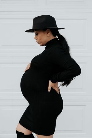 Maternity fashion portrait in black dress and wide-brim hat against white background.