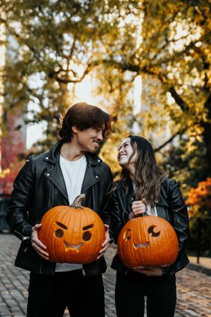 Couple in leather jackets holding carved Halloween pumpkins during autumn sunset.