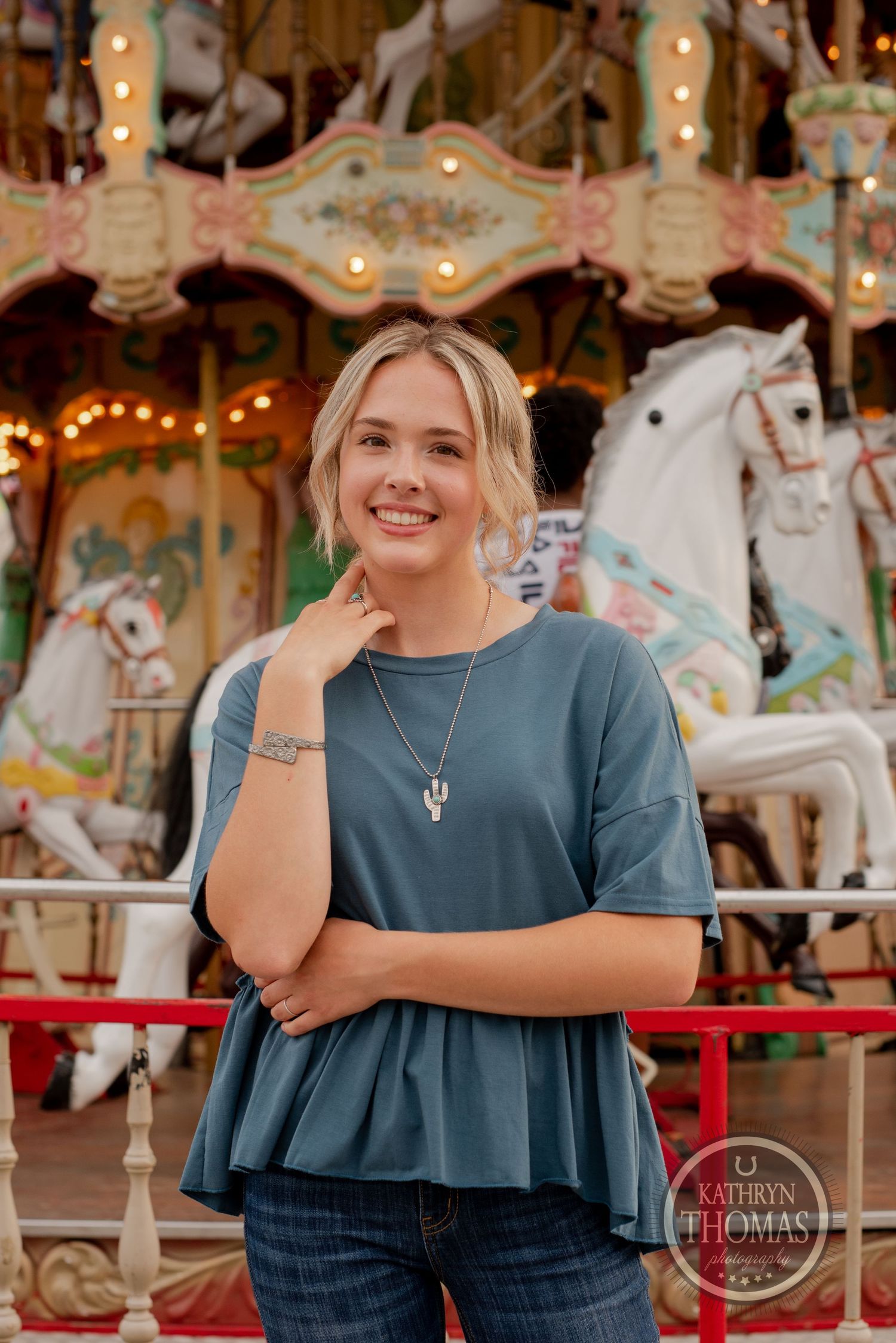 Person wearing teal peplum top poses in front of illuminated carousel horses at carnival.