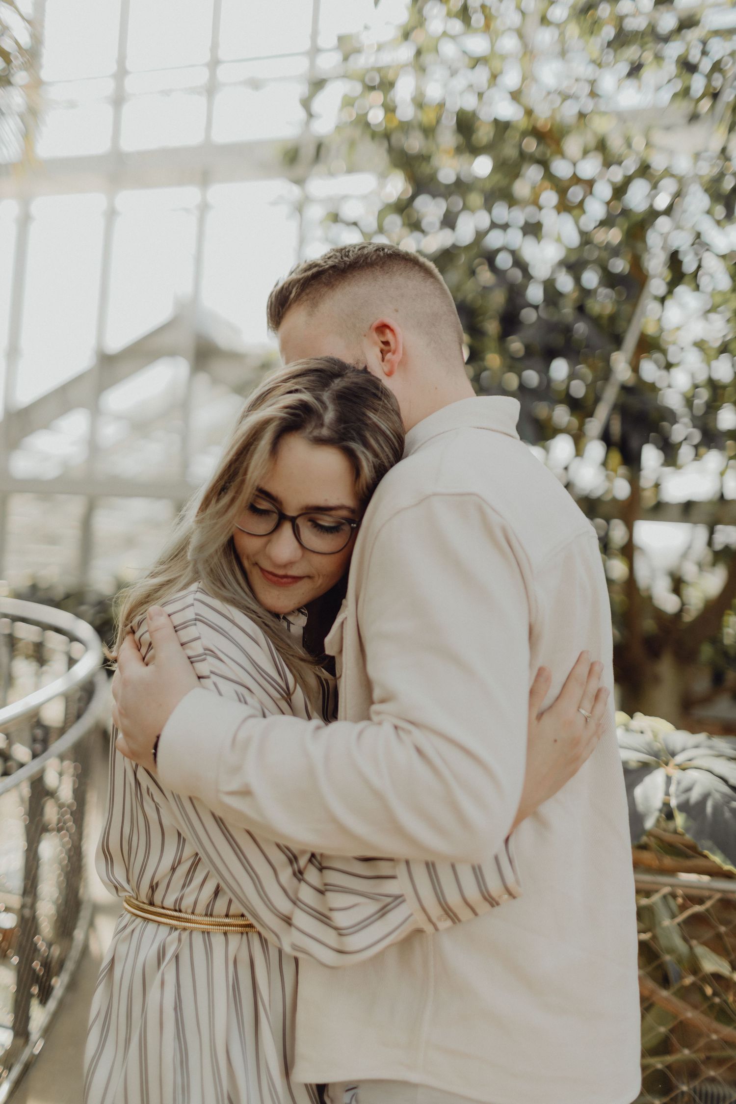 Intimate couple embrace in light-filled greenhouse wearing neutral toned clothing.
