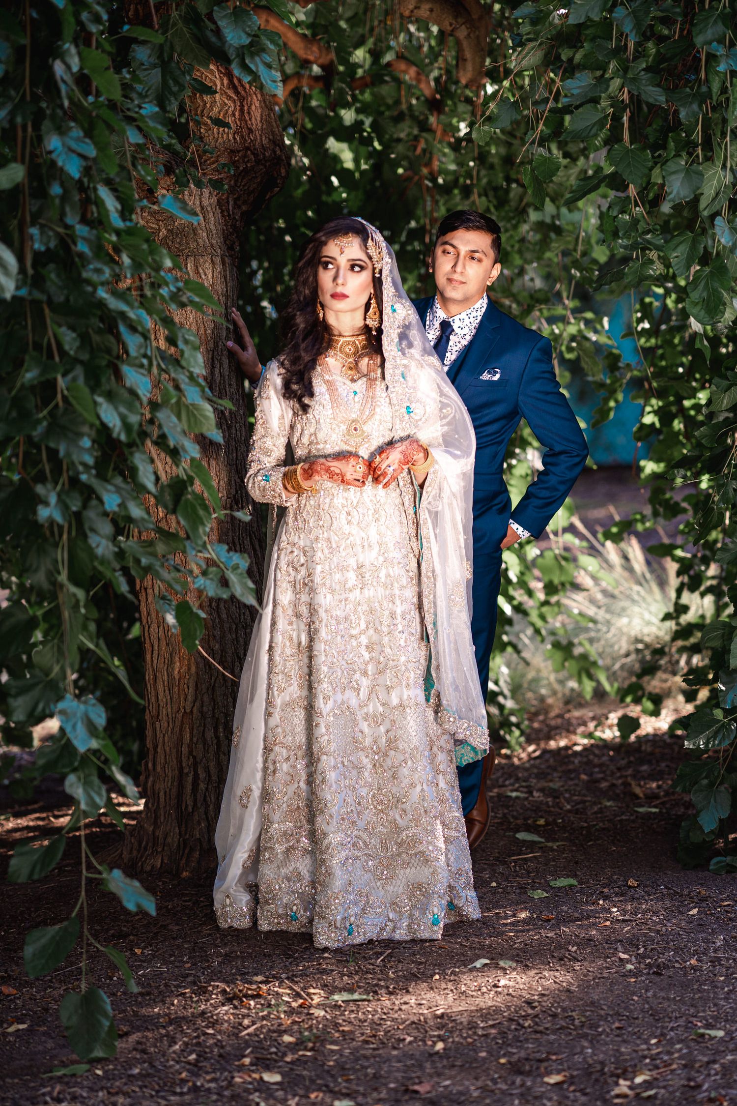 Pakistani groom peeks from behind, under a dark green tree, sharing a secret gaze with his bride, love hidden in foliage