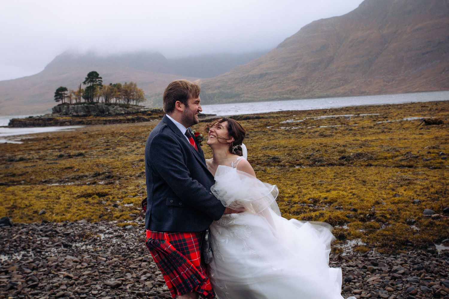 Wedding couple embracing on rocky shore of Scottish loch with mountains in background.