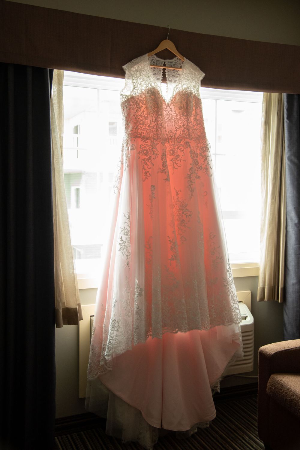 White lace wedding dress with delicate details hanging in window light.