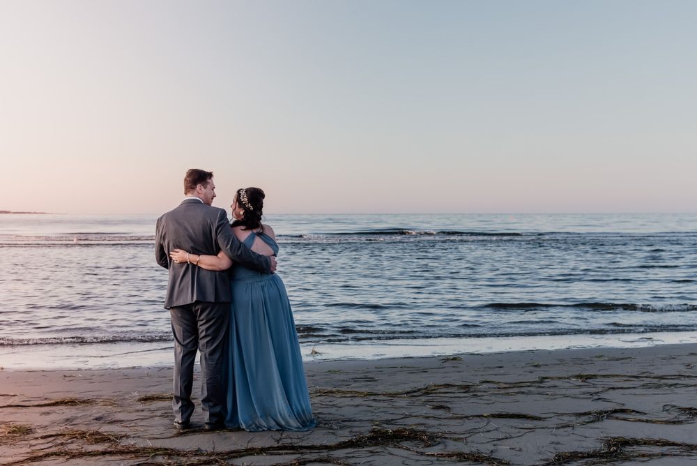 Couple embraces on beach at sunset wearing formal blue dress and grey suit.
