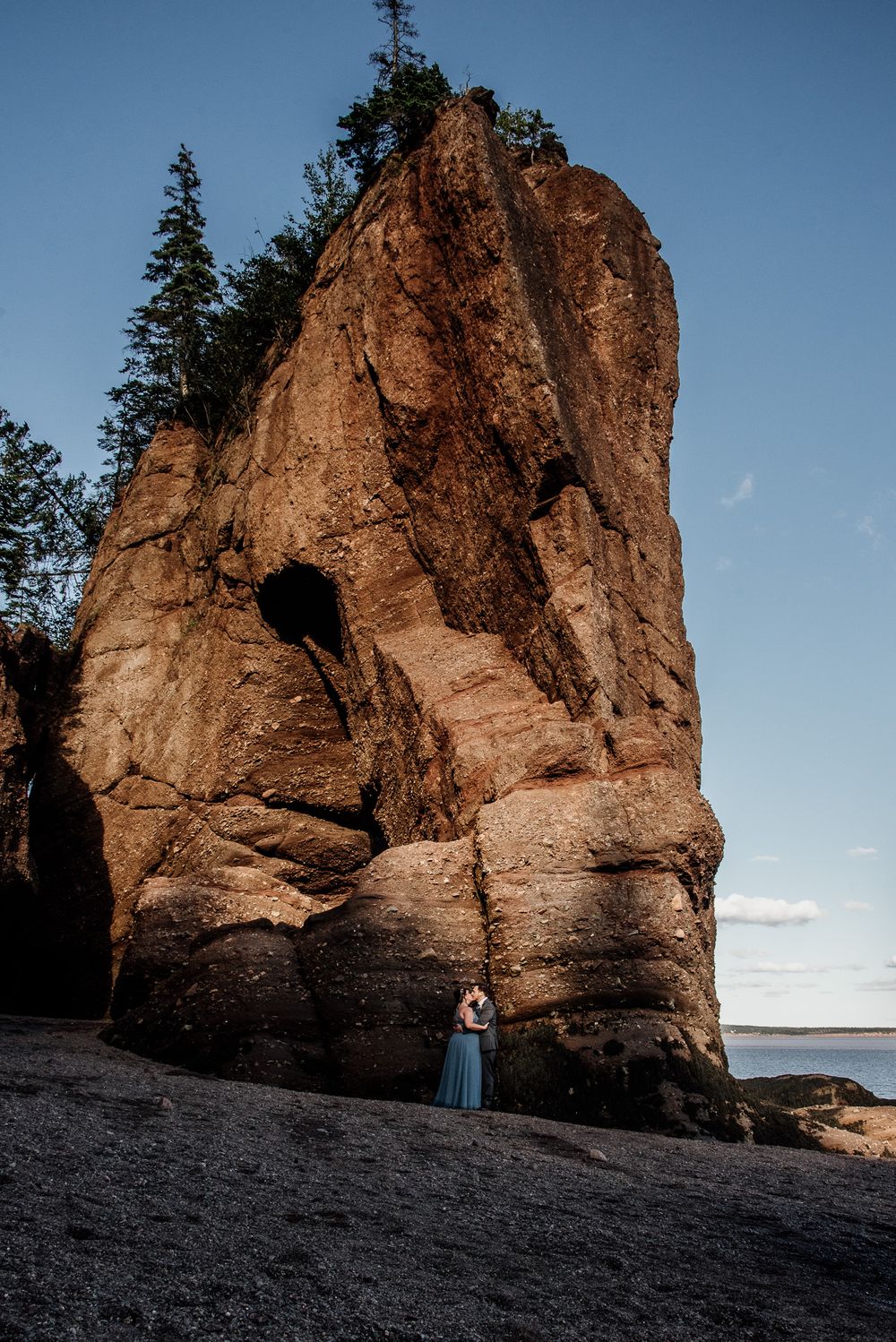 Couple in wedding attire stand beneath towering coastal rock formations at sunset.