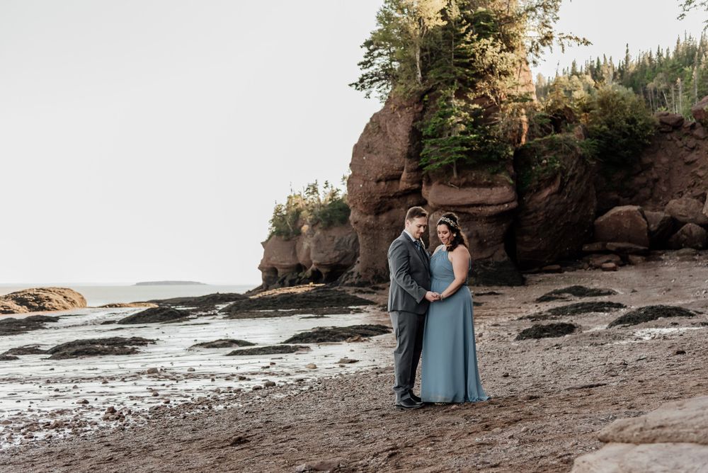 Couple in formal wear embracing on rocky beach with dramatic cliffs in background.