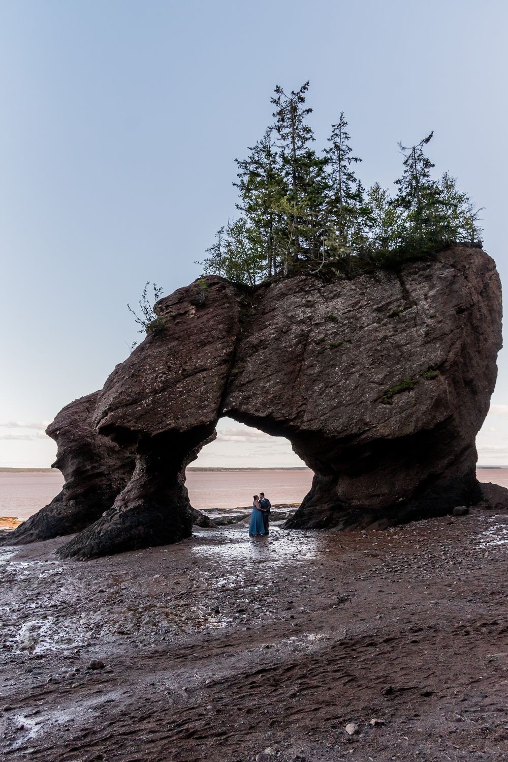 Couple standing under natural rock arch formation at low tide on beach.