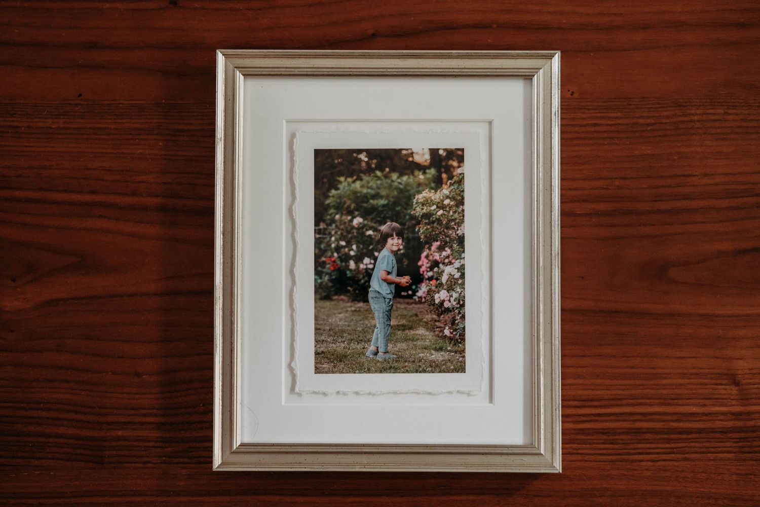 A framed vintage photograph with deckled edge showing a figure in light blue clothing in a garden setting, mounted on a wooden wall.