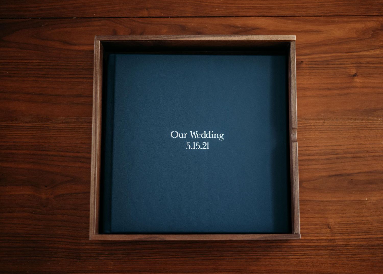 A dark blue wedding album with the text 'Our Wedding 5.15.21' in a custom walnut album box centered on a wooden surface.