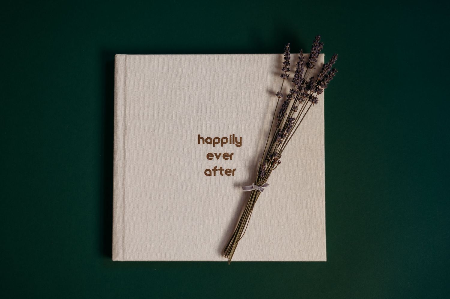 White linen wedding photo album with 'happily ever after' text on cover and dried flowers against dark green background.