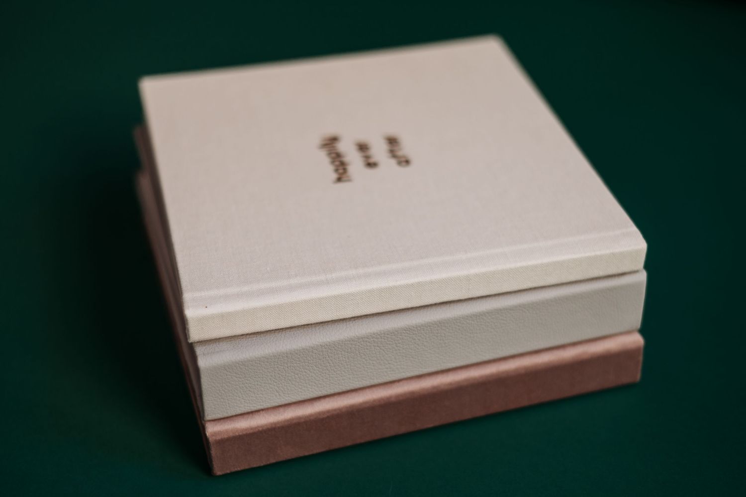 A white square wedding album with soft metallic lettering sits on a dark green surface.