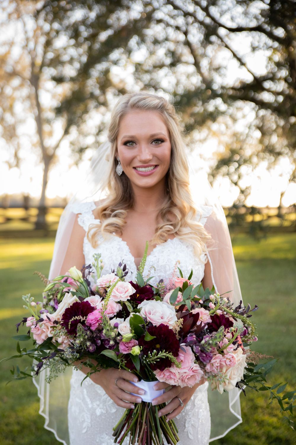 Smiling bride in lace gown holds lush purple and pink floral bouquet at golden hour.