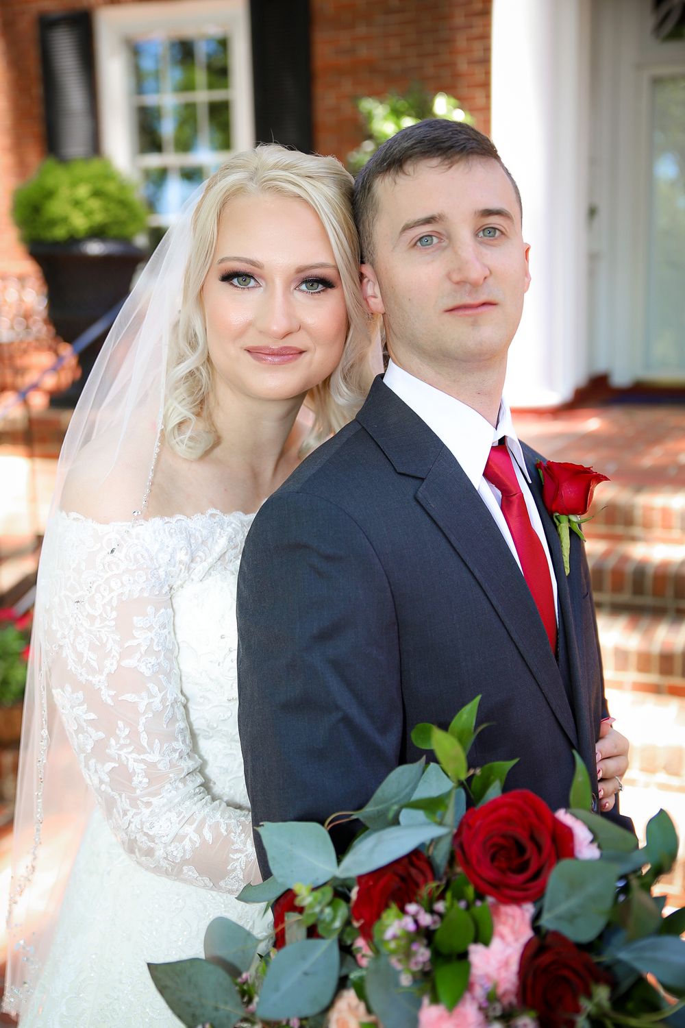 Bride and groom pose together outside brick building with red roses bouquet.