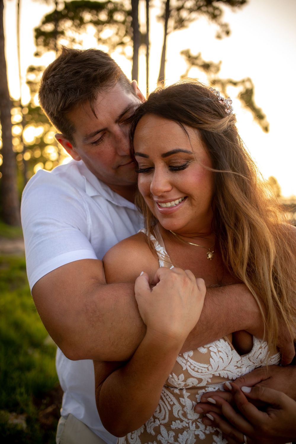 Romantic couple embracing at sunset outdoors, woman in lace dress smiling as man holds her from behind near trees.
