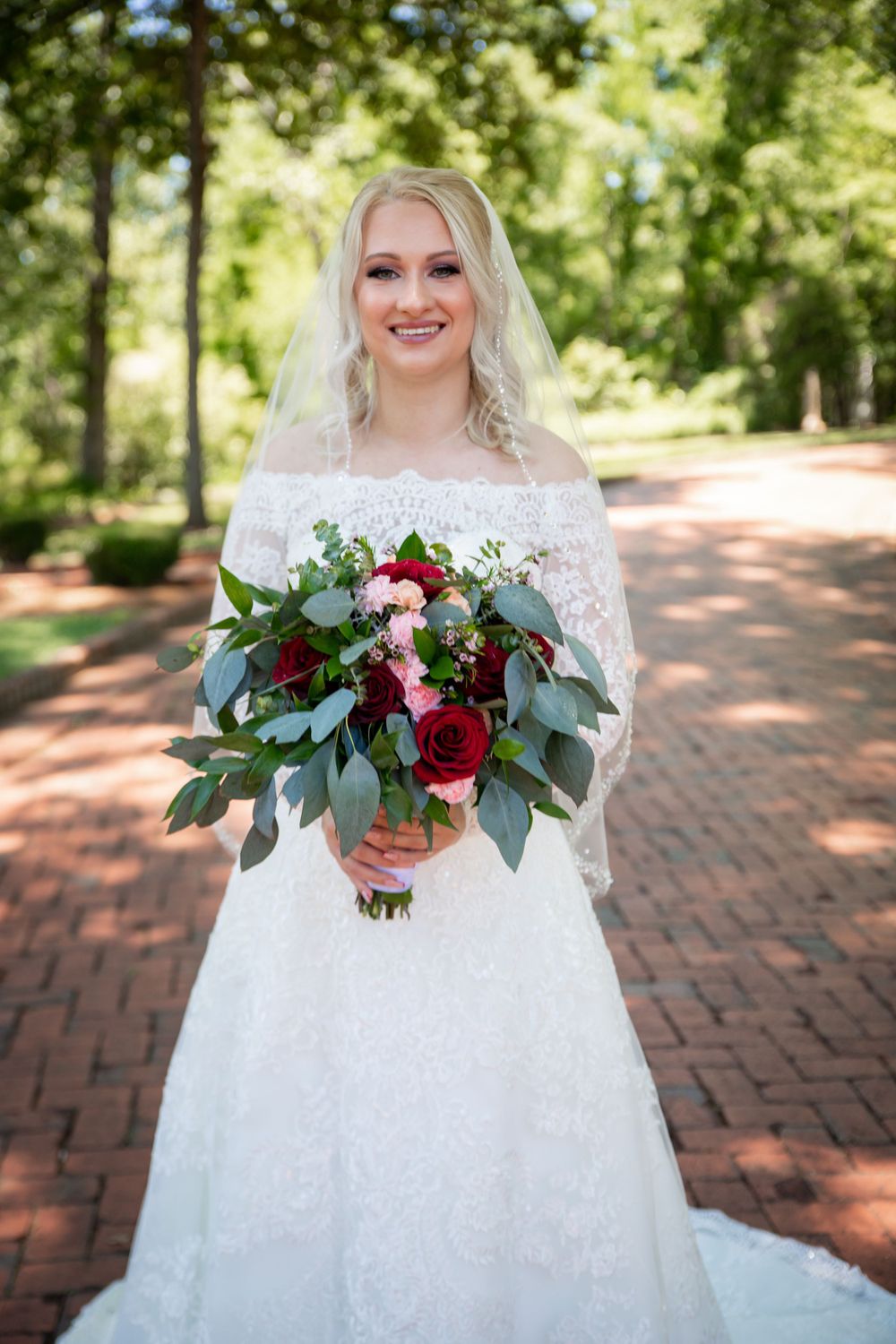 Blonde bride in lace off-shoulder gown holds red and pink bouquet on brick pathway.