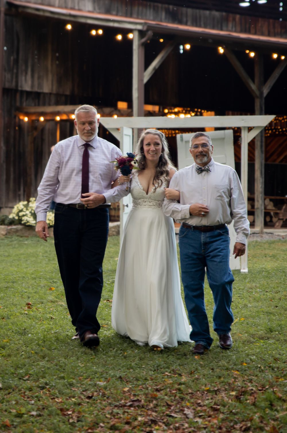 Bride walks down the aisle escorted by two men at a rustic barn wedding venue.
