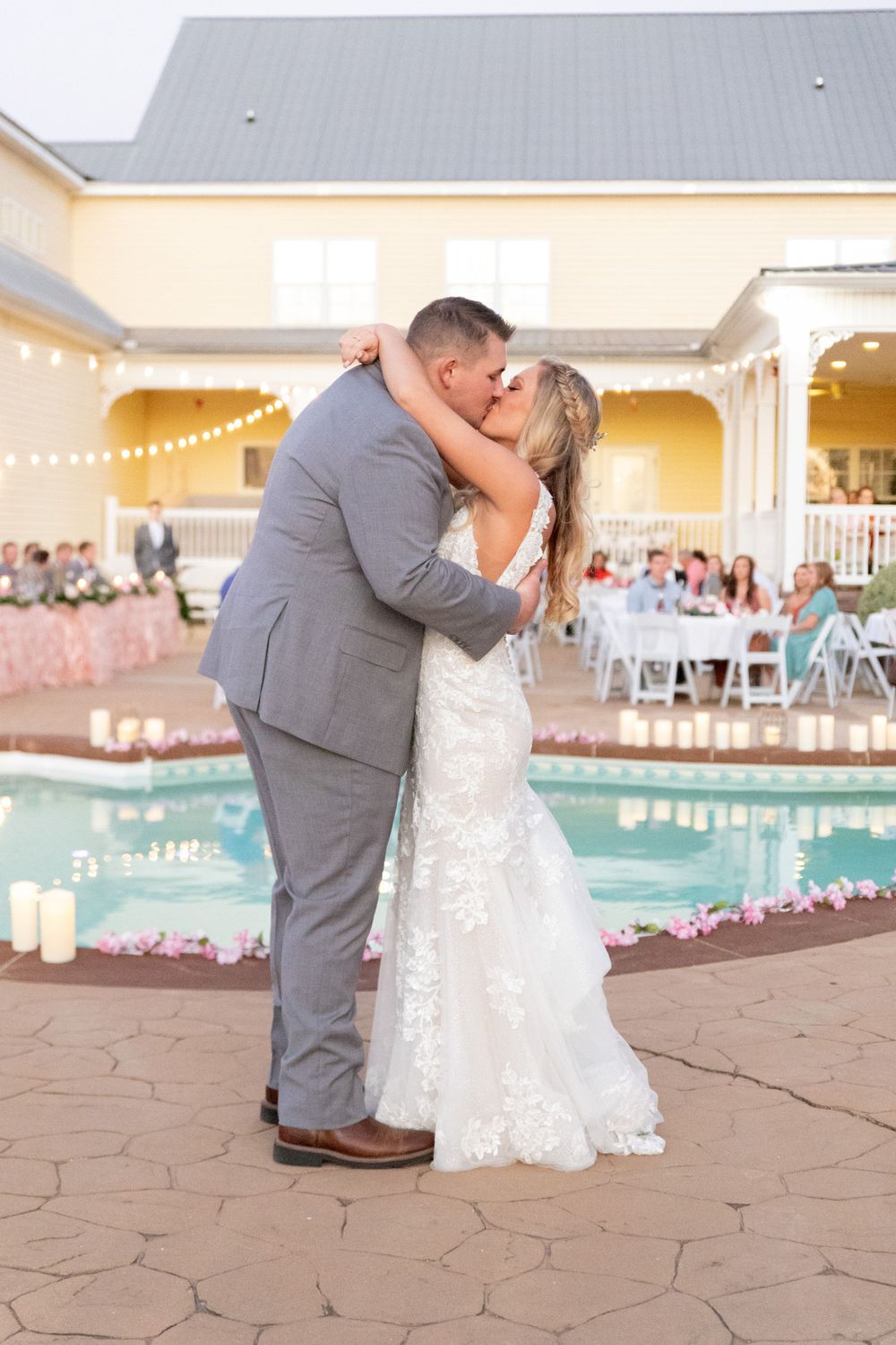 Bride and groom share first dance kiss beside flower-lined pool at elegant outdoor wedding reception at dusk.