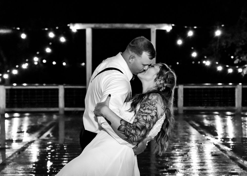 Black and white romantic photo of bride and groom kissing in the rain under twinkling lights at night.