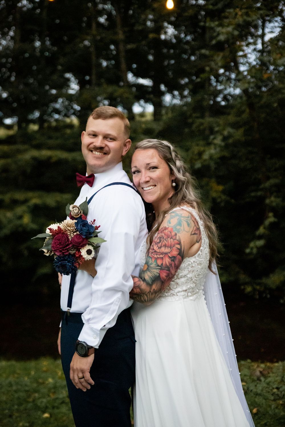 Smiling bride with floral tattoo sleeve leans against groom in white shirt with suspenders holding bouquet outdoors.