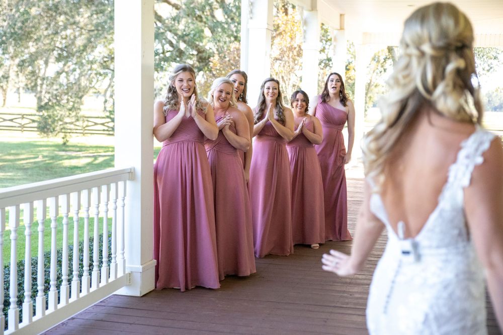 Bridesmaids in pink dresses react with joy on a porch as bride reveals her wedding gown.