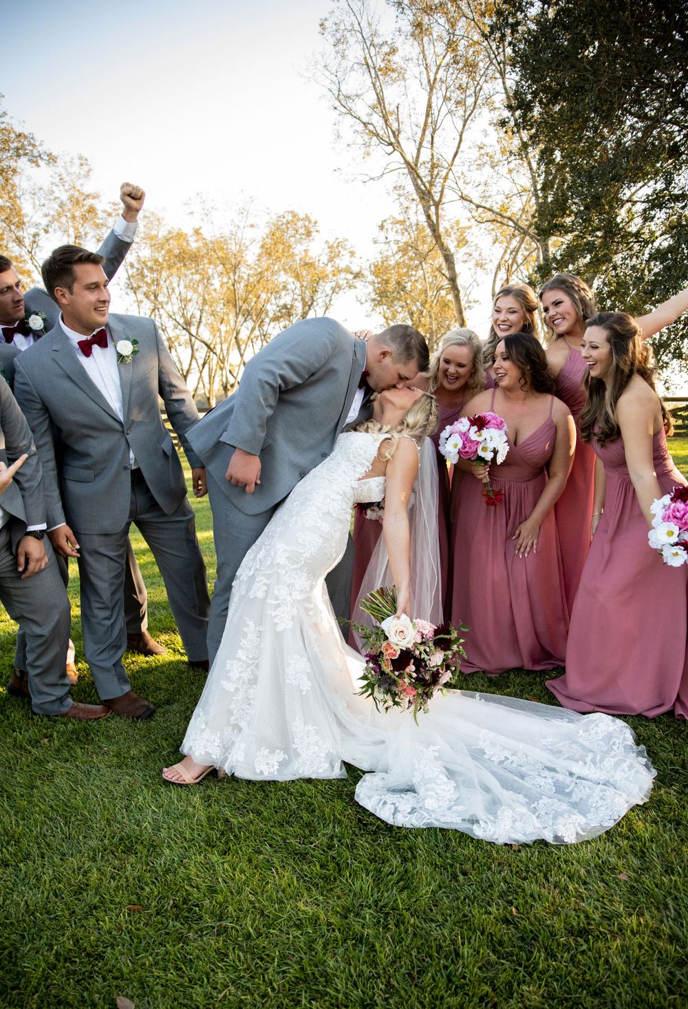 Groom dipping bride for a kiss surrounded by cheering wedding party in gray suits and dusty rose bridesmaid dresses.