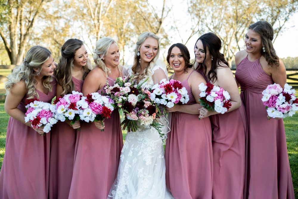 Bride in lace gown laughing with five bridesmaids in dusty rose dresses holding pink and white bouquets outdoors.