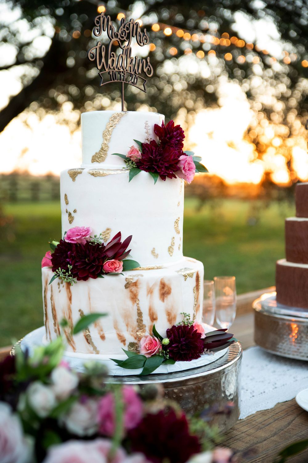Elegant three-tier white wedding cake with gold accents and burgundy floral decorations at outdoor sunset reception.