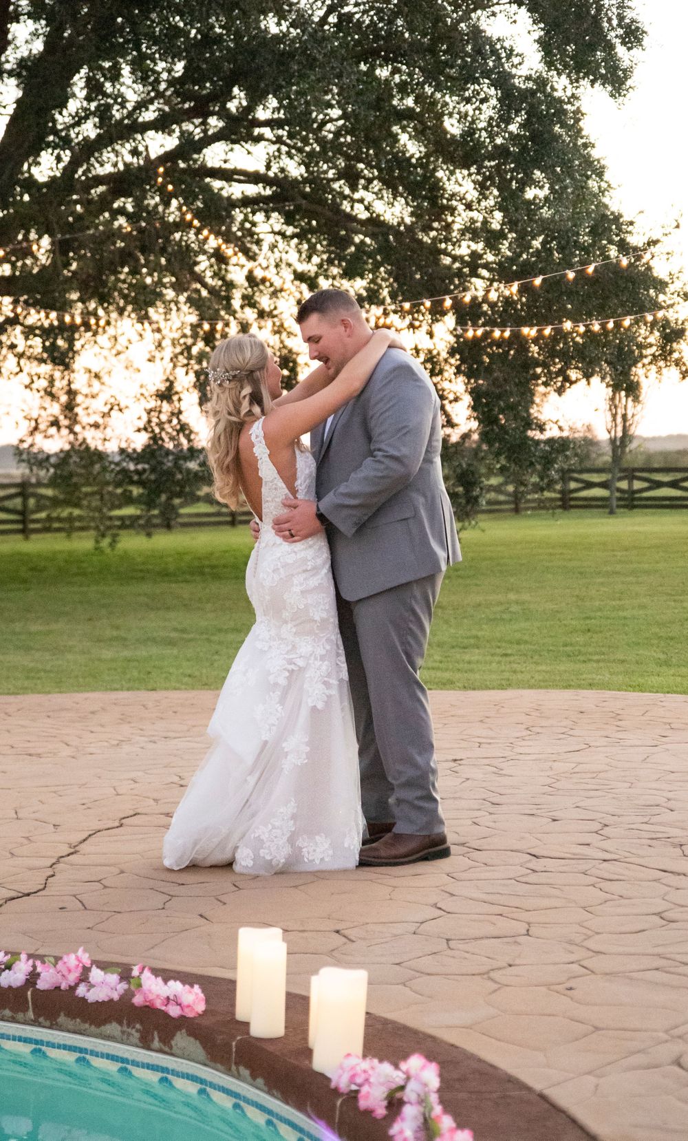 Bride and groom share first dance on stone patio by candlelit pool at sunset wedding reception.