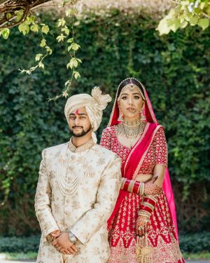Traditional Indian wedding portrait with couple in red lehenga and cream sherwani against garden backdrop.