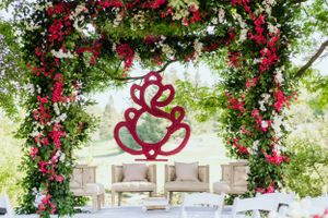 An outdoor wedding arch decorated with red roses and greenery features a stylized heart design.