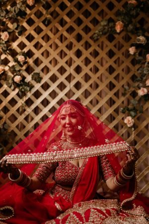 A joyful bride in red traditional attire poses with her dupatta against a floral lattice backdrop.