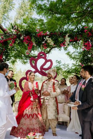 Outdoor Indian wedding ceremony with guests gathered under floral arch and red heart decoration.