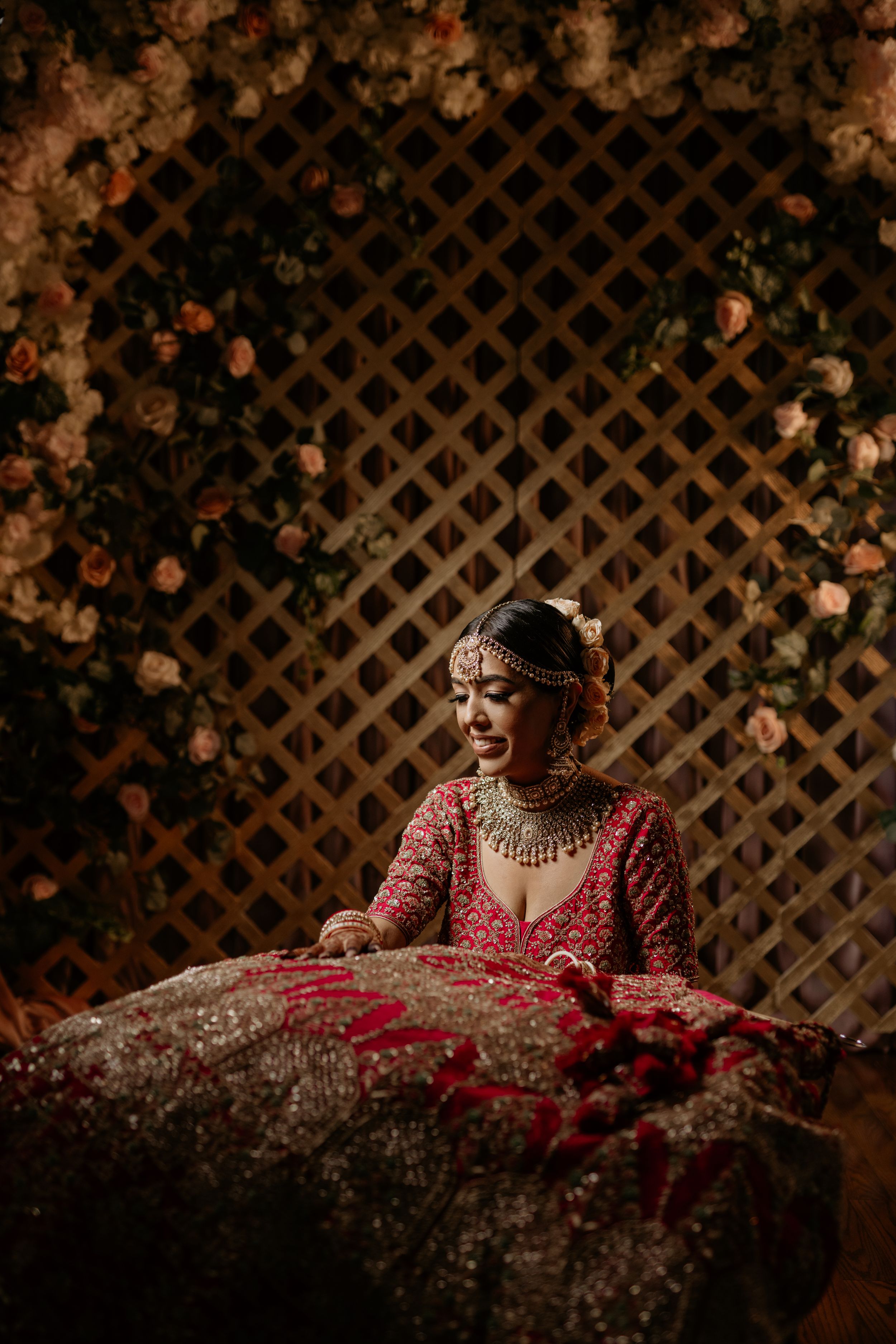 A traditional South Asian bride wearing an ornate red lehenga poses against a lattice backdrop with flowers.