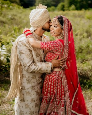 A traditional South Asian wedding couple embraces in ornate red and beige wedding attire outdoors.