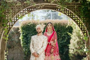 Couple in traditional Indian wedding attire posing under decorative garden arch.