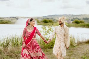 Bride in red lehenga and groom in cream sherwani photographed by lakeside.