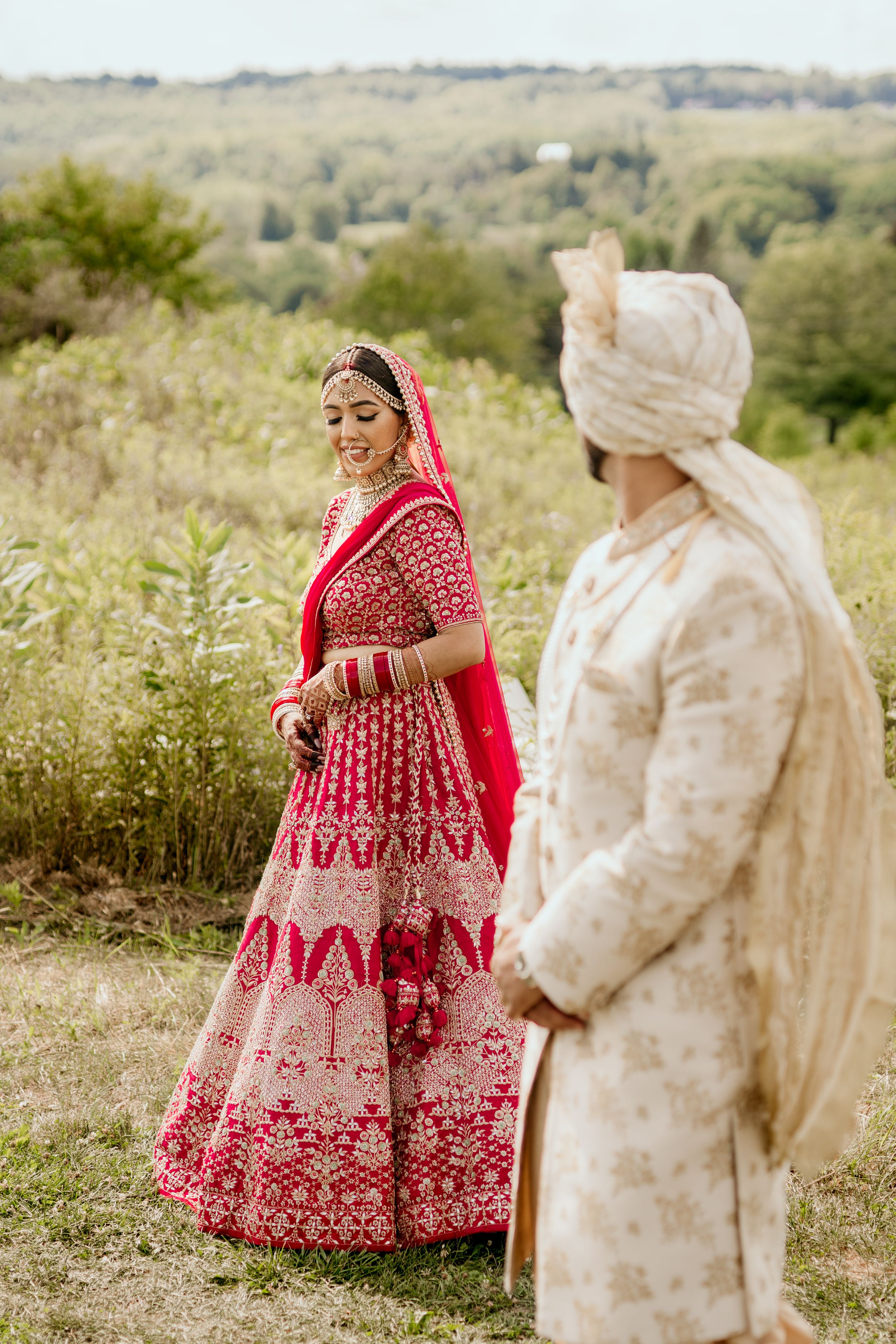 A bride in a red lehenga poses elegantly while the groom looks on in a countryside setting.