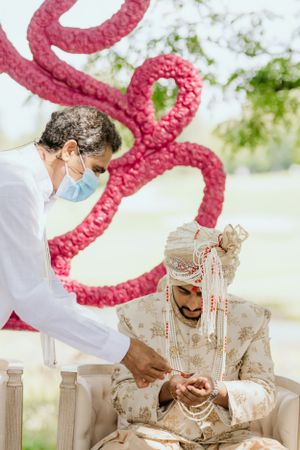 Wedding ceremony with protective masks and traditional Indian attire under pink decorative arch.