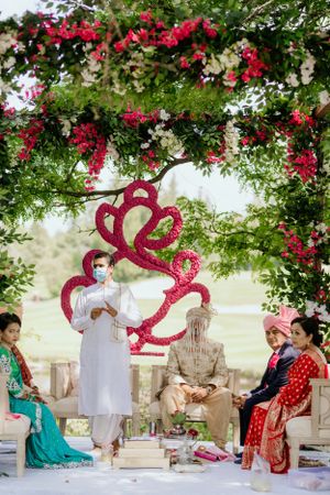 Outdoor Indian wedding ceremony under arch with red floral decorations and pink heart design.