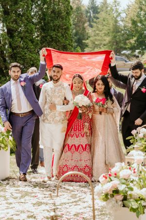 Wedding party holds red fabric canopy while walking down garden pathway.