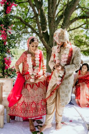 Traditional Indian wedding ceremony under flowering trees with couple in red and beige formal attire during jaimala ritual.