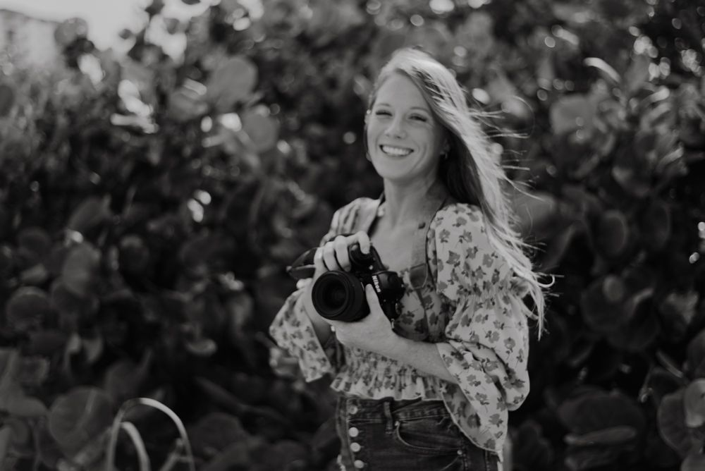 Photographer with long hair holding camera outdoors in black and white.