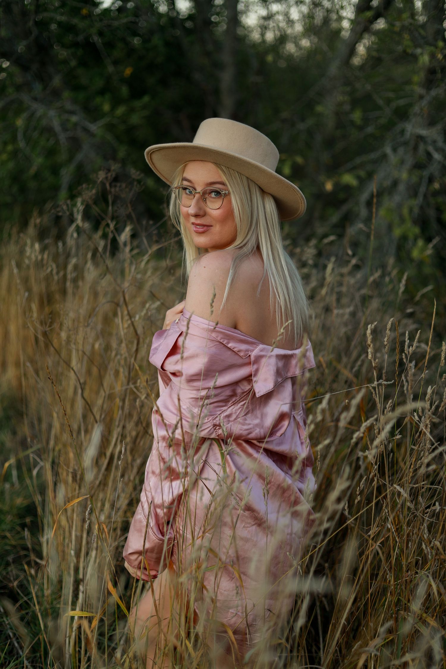 A dreamy portrait in tall grass features a pink off-shoulder dress and wide-brimmed hat with glasses.