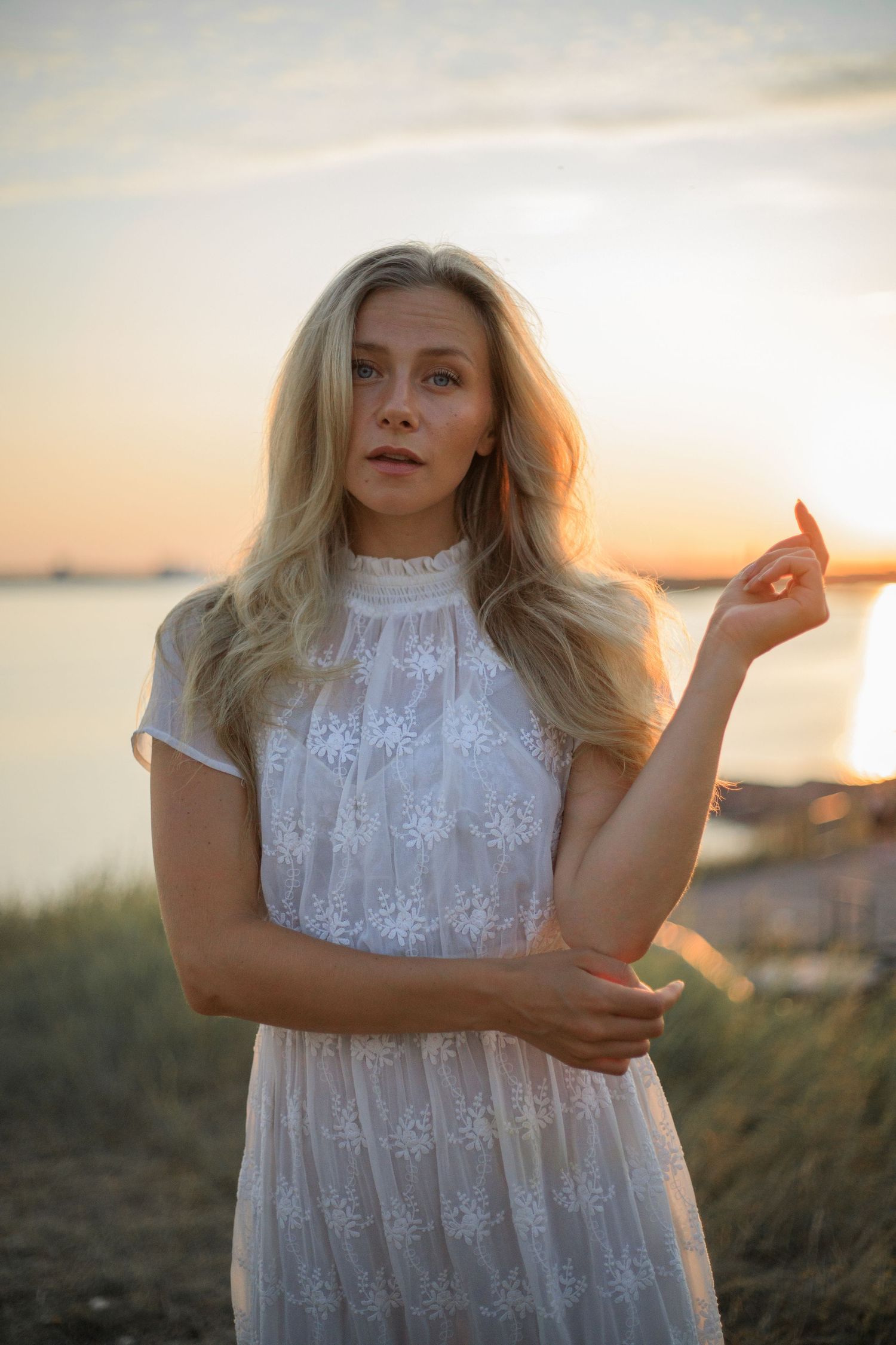 Woman in white lace dress posing at sunset by the water.
