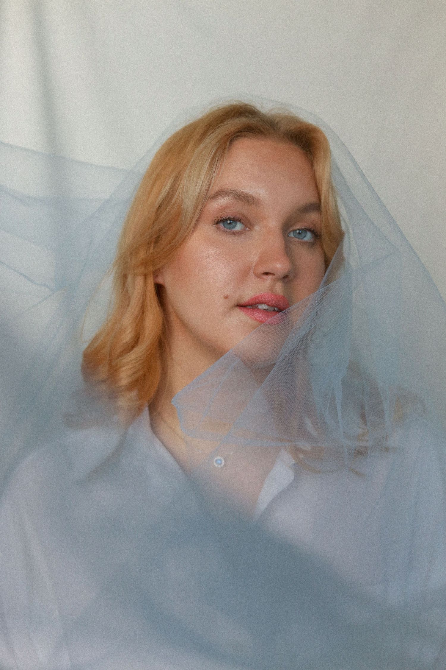 Artistic portrait through sheer blue fabric creates dreamy ethereal effect with soft natural lighting.