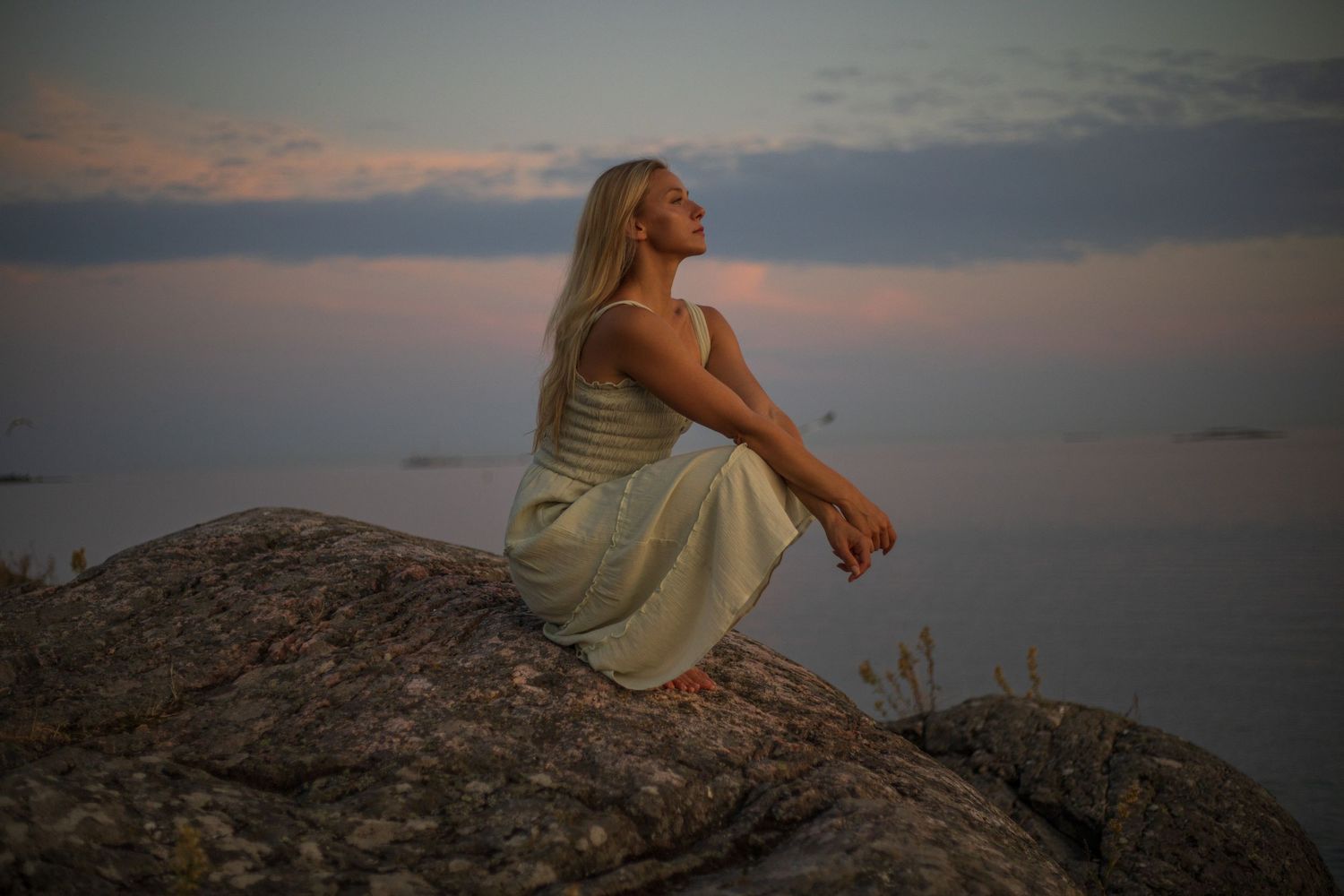 A person in a dress sits on a rocky cliff overlooking a peaceful ocean view at sunset.