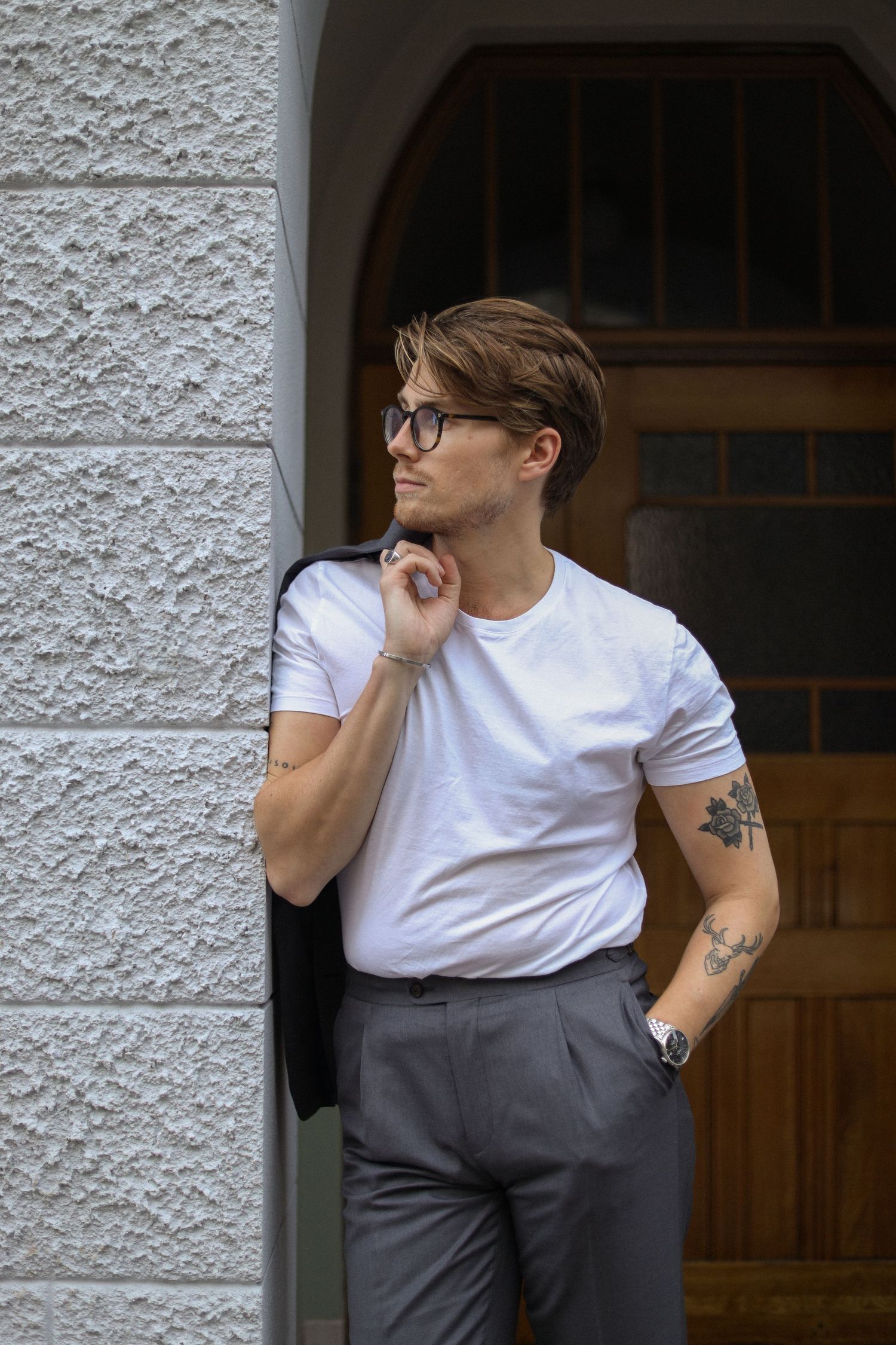 A person in a white t-shirt and grey pants leans against a textured wall, hand touching neck thoughtfully.