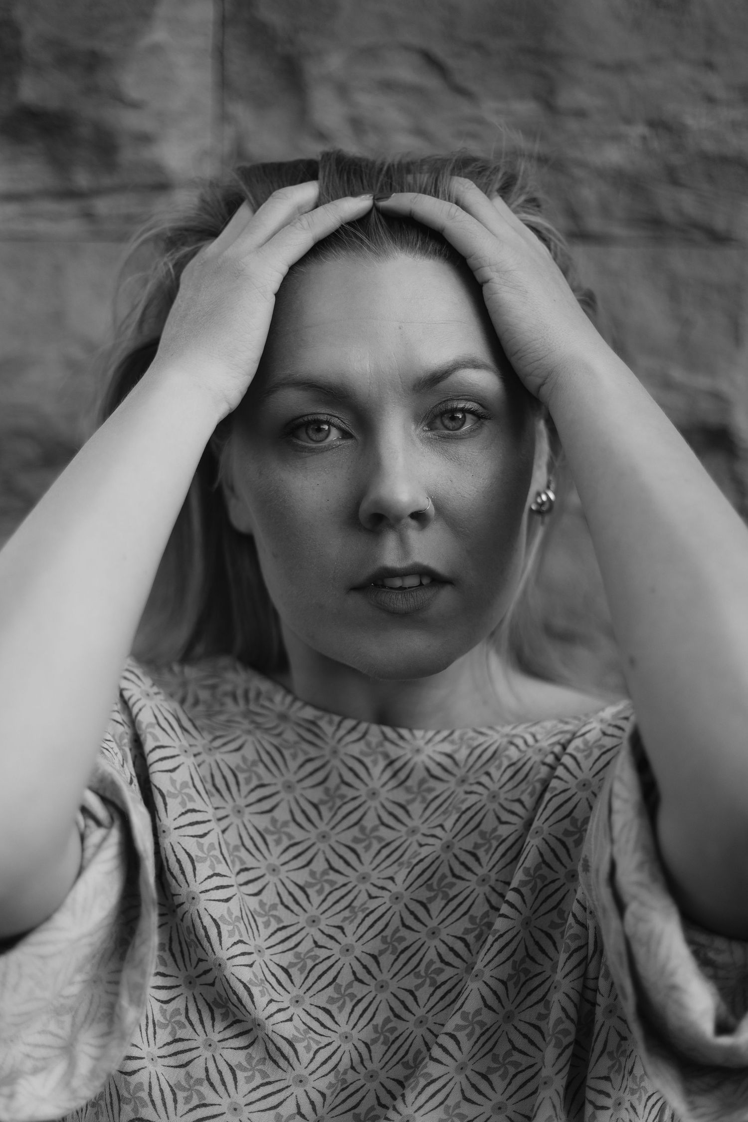 Black and white portrait with hands placed on head against a textured wall.