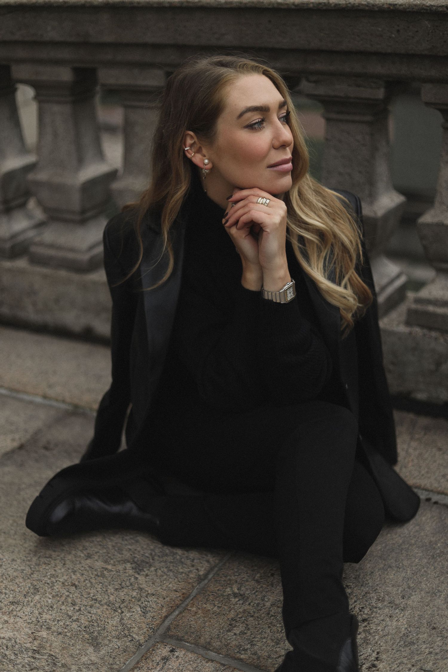 Fashion model in all black attire sits thoughtfully on stone steps in moody outdoor setting.