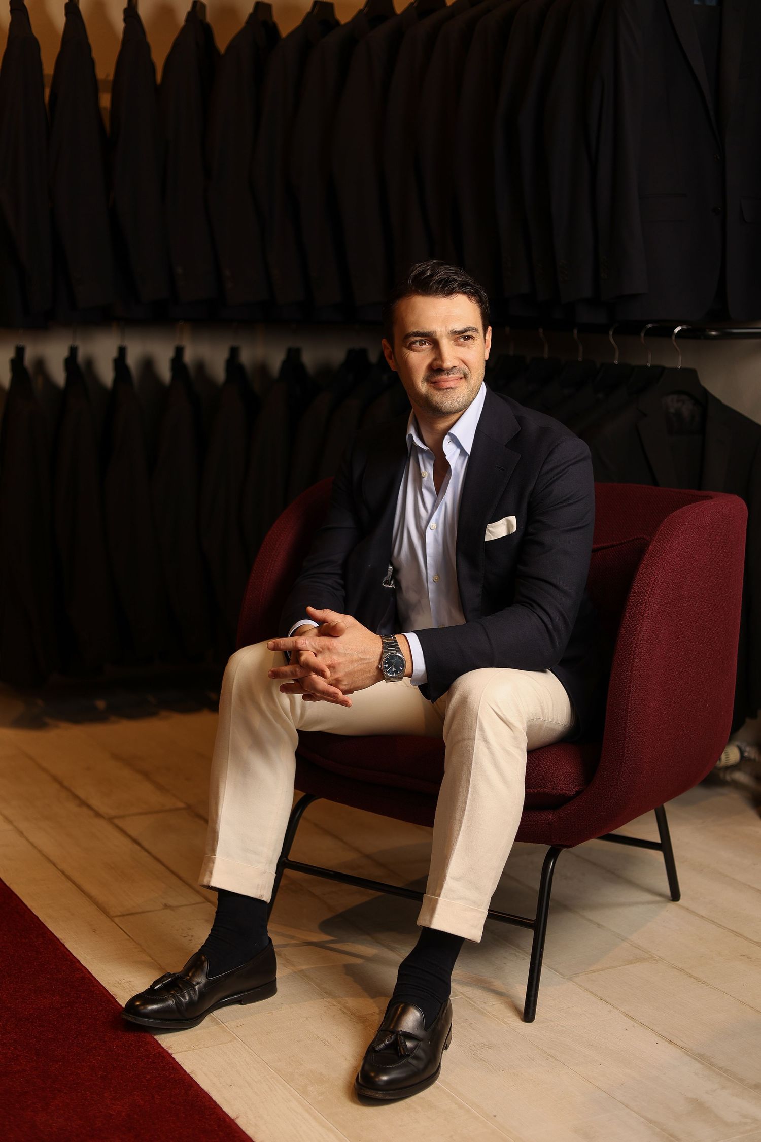 Well-dressed man sitting in modern chair against dark wall with suits.